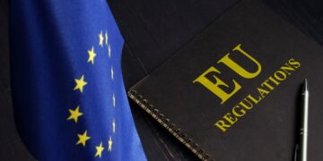 A small European Union flag with yellow stars on a blue background is on the left. On the right, a black notebook with "EU Regulations" in bold yellow text sits on a dark surface, hinting at updates from EUdigitallaw.com. A silver pen lies beside the notebook.