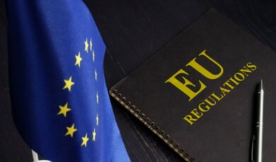 A small European Union flag with yellow stars on a blue background is on the left. On the right, a black notebook with "EU Regulations" in bold yellow text sits on a dark surface, hinting at updates from EUdigitallaw.com. A silver pen lies beside the notebook.